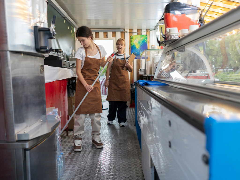 Two employees in brown aprons inside a food truck, one mopping the floor while the other holds a clipboard, illustrating a commercial cleaning company in Marietta.