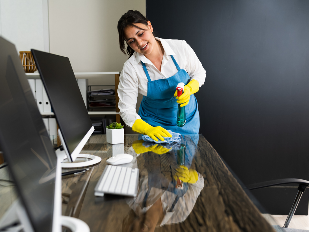 A person in a blue apron and yellow gloves wipes a glass office desk with a spray bottle and cloth, representative of office cleaning cost in Marietta.