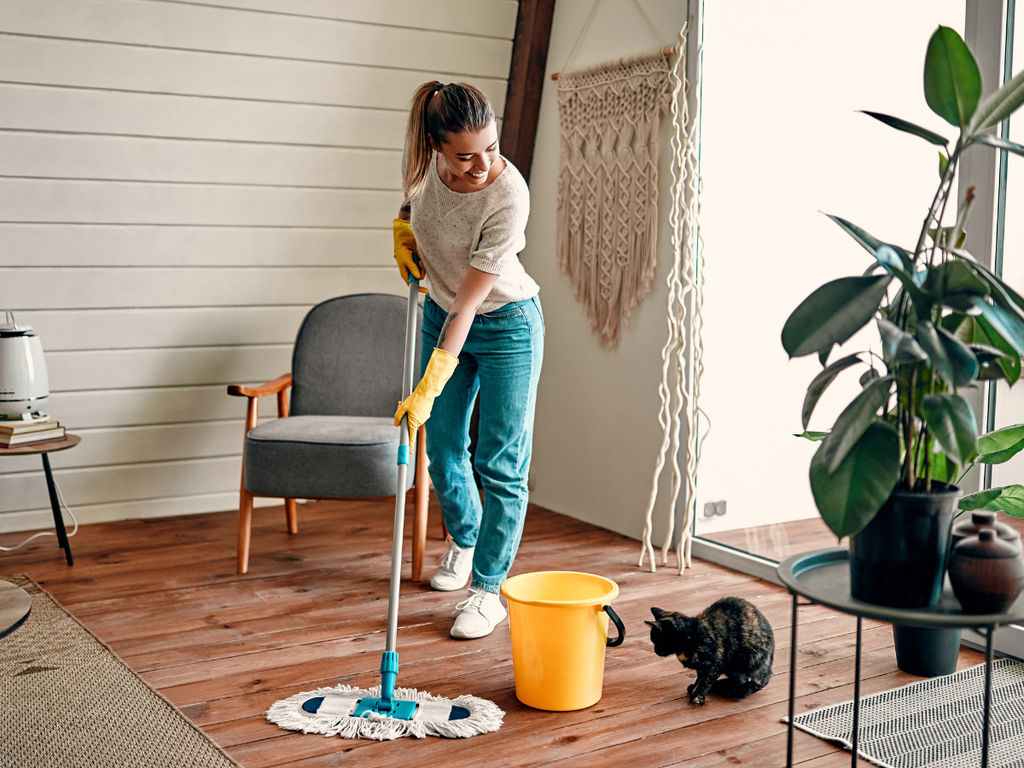 A woman in yellow rubber gloves mops a wooden floor next to a cat and a bucket, a common task to consider when reviewing questions to ask a house cleaning service.