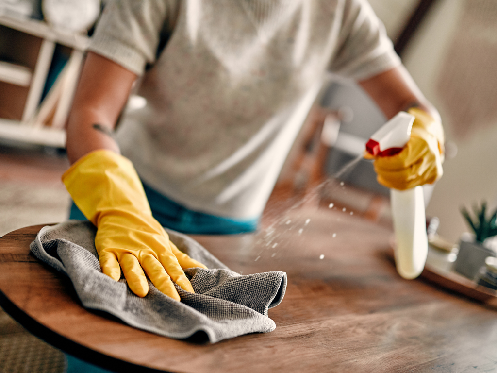 Person wearing yellow rubber gloves cleaning a wooden table with a spray bottle and a cloth, the kind of surface maintenance mentioned in questions to ask a house cleaning service.
