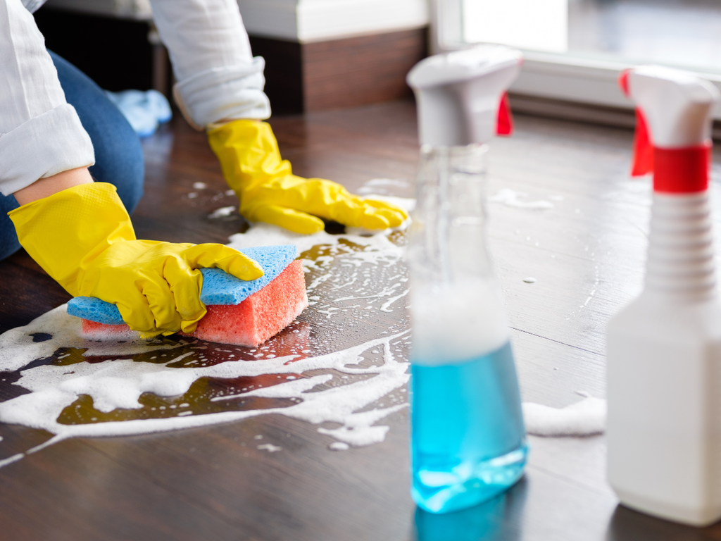 A person wearing yellow rubber gloves scrubs a soapy wooden floor, a task relevant when considering questions to ask a house cleaning service, with two spray bottles in the foreground.