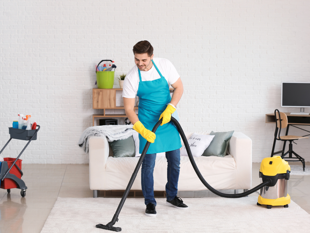 A man in an apron and yellow gloves vacuums a white rug in a living room, a task often discussed when reviewing questions to ask a house cleaning service.