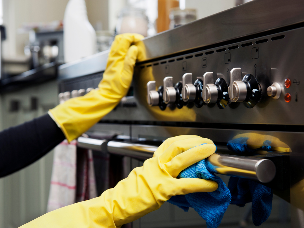 A person wearing yellow rubber gloves cleaning a stainless steel oven, highlighting the level of detail to look for when researching questions to ask a house cleaning service.