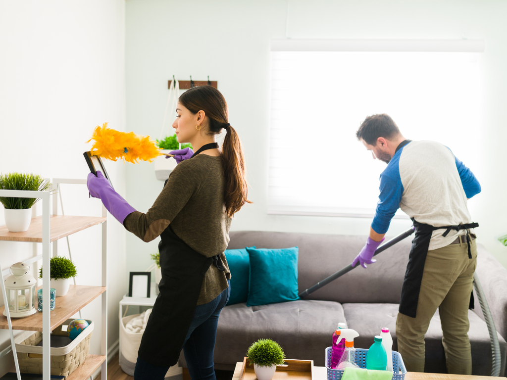 A woman uses a feather duster on a picture frame as a man vacuums a gray sofa, a scene that often prompts questions to ask a house cleaning service.